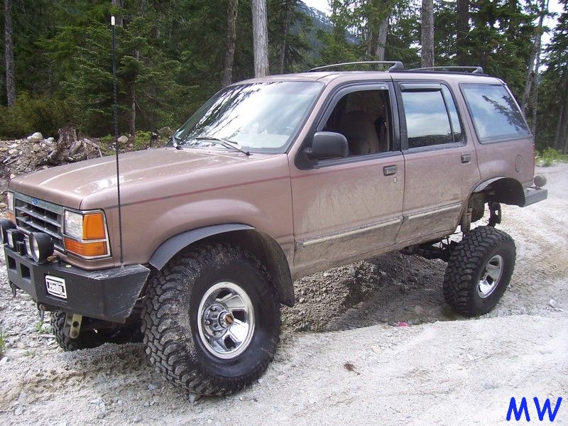 Lifted tan SUV with large off-road tires and custom bumper in a forest setting.