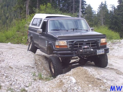 Lifted dark pickup truck with white camper shell on rocky terrain, front wheel elevated.