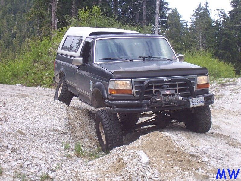 Lifted dark pickup truck with white camper shell on rocky terrain, front wheel elevated.