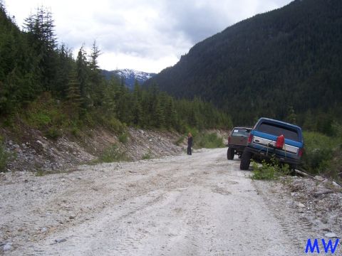 Blue Ford Bronco parked on a gravel road in a forest with mountains in the background.
