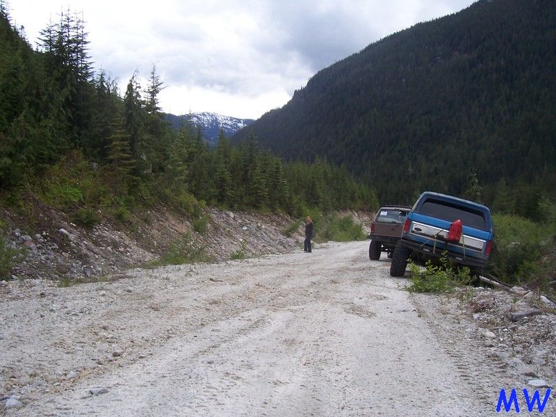 Blue Ford Bronco parked on a gravel road in a forest with mountains in the background.