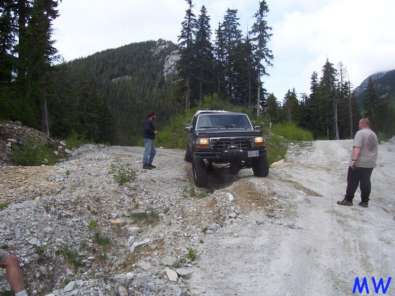Dark Ford Bronco driving up a rocky mountain road with two men nearby.
