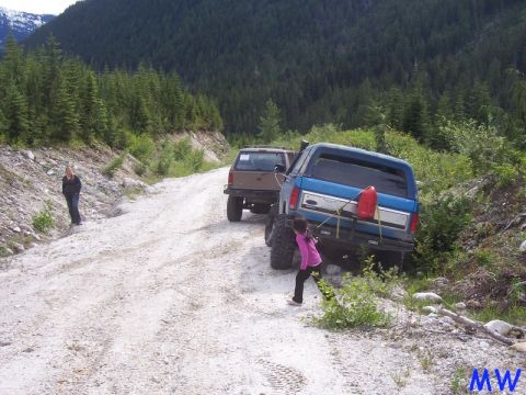 Blue Ford Bronco with red gas can on rear bumper on a dirt road, girl walking past.