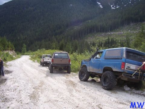 Blue lifted Ford Bronco on a gravel road with forest and mountains in the background.