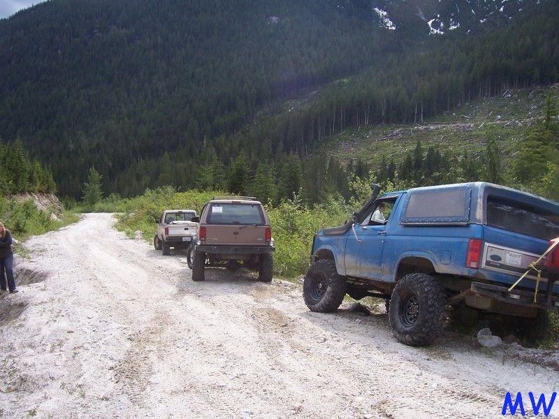 Blue lifted Ford Bronco on a gravel road with forest and mountains in the background.