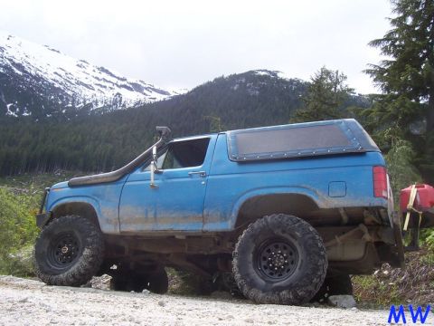 Blue 1980s Ford Bronco with snorkel and off-road tires on a mountain road.