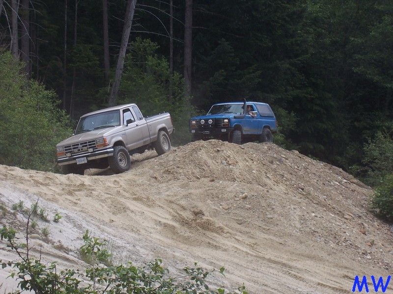 Tan pickup truck and blue SUV off-roading on a steep dirt hill with trees in the background.