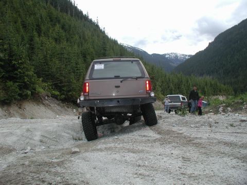 Lifted brown Ford Explorer on a rocky off-road trail with people and other vehicles in the background.