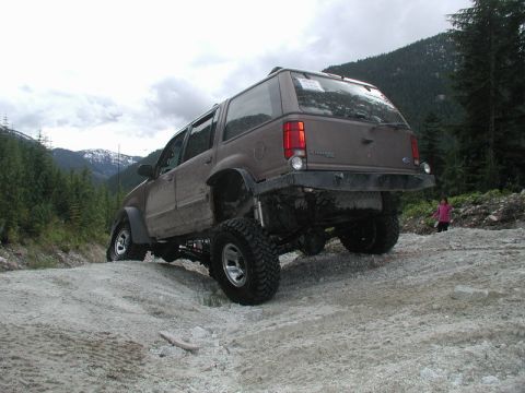Lifted brown Ford Explorer off-roading on a gravel hill with mountains and trees.