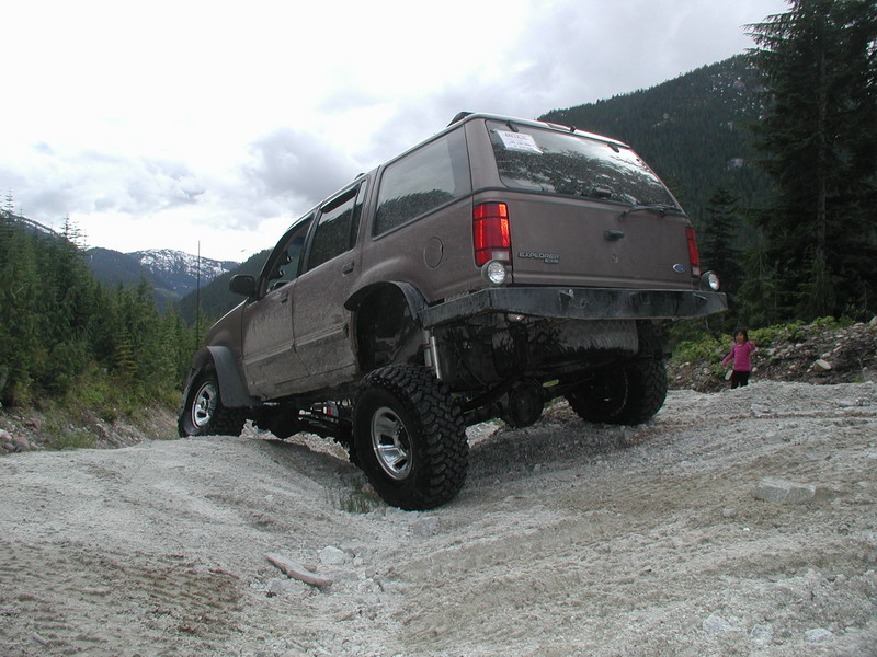 Lifted brown Ford Explorer off-roading on a gravel hill with mountains and trees.