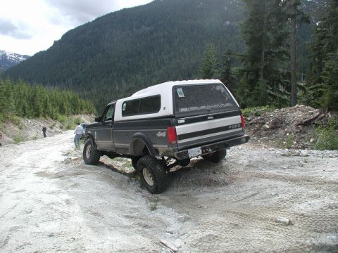 Lifted Ford pickup truck with white camper shell on a dirt road in a mountainous forest.