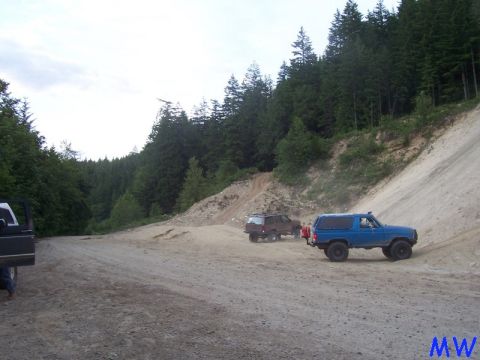 Blue Ford Bronco parked on a dirt road with a sandy hill and forest.
