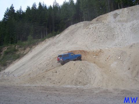Blue Ford Bronco driving up a steep, sandy hill with trees in the background.