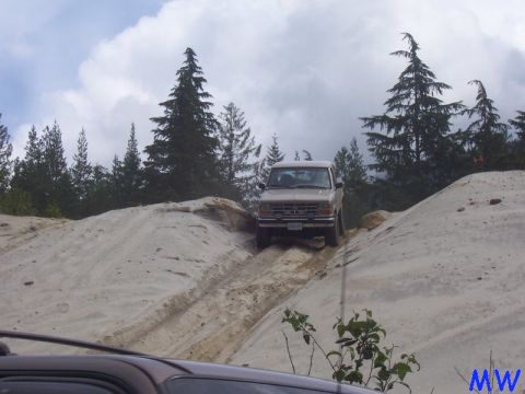 Tan 1980s Ford Bronco driving up a sandy hill with trees in the background.
