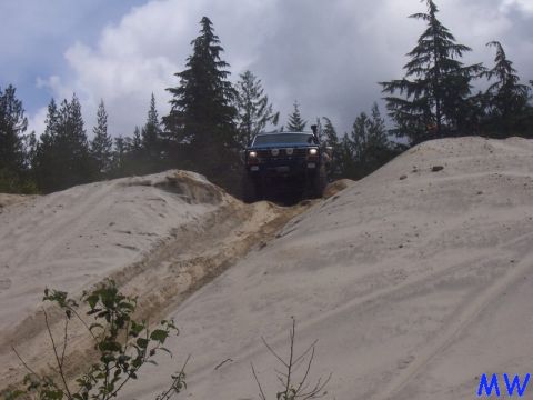 1985 Ford Bronco driving up a sandy hill with trees in the background.