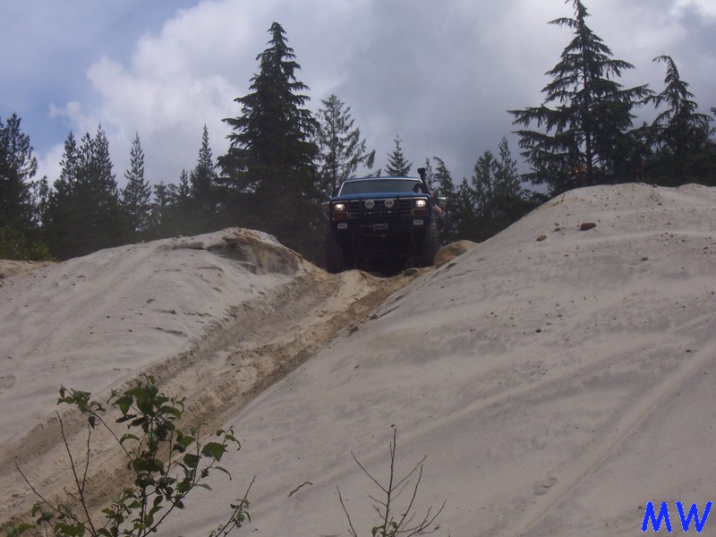 1985 Ford Bronco driving up a sandy hill with trees in the background.