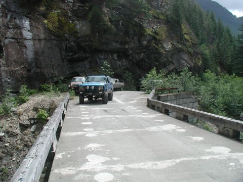 Blue Ford Bronco on a bridge with other vehicles in a mountain forest.