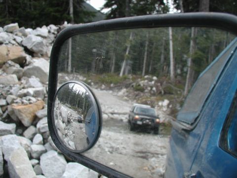 Reflection in a blue truck's side mirror shows another vehicle on a rocky, forested off-road path.