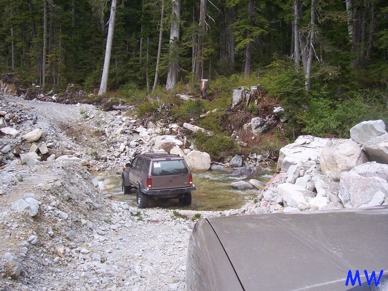 Brown SUV driving through a stream on a rocky forest road.