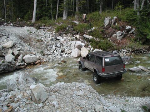 Brown SUV driving through a rocky stream in a forest.