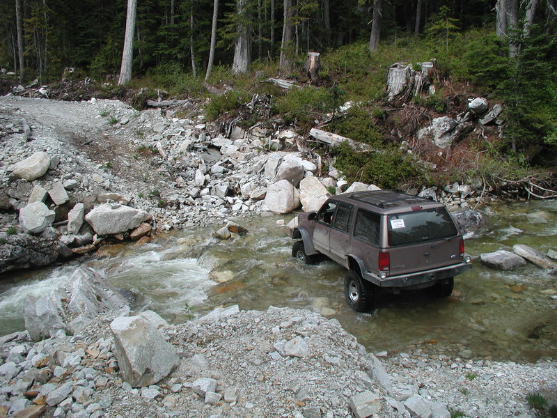 Brown SUV driving through a rocky stream in a forest.
