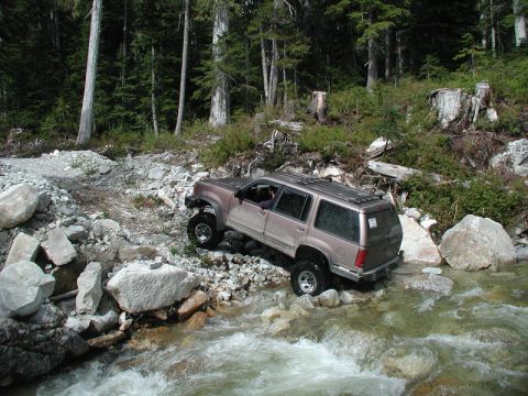 Tan SUV navigating a rocky stream in a forest.