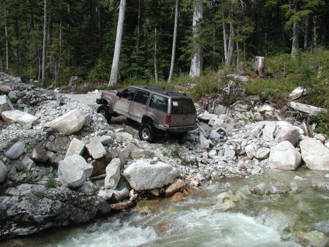 Tan SUV driving on a rocky trail next to a stream in a forest.