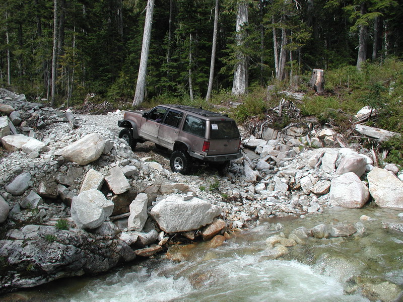Tan SUV driving on a rocky trail next to a stream in a forest.