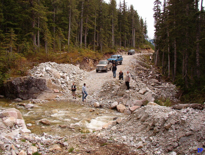 People and off-road vehicles at a rocky stream crossing on a dirt road in a forest.