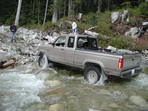 Tan Ford pickup truck driving through a rocky stream with two people nearby.