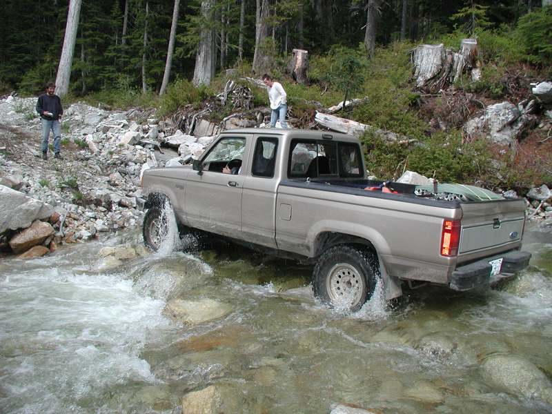 Tan Ford pickup truck driving through a rocky stream with two people nearby.