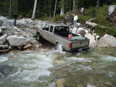 Tan Ford pickup truck driving through a rocky stream, with trees and boulders on the bank.