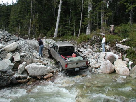 Tan Ford pickup truck driving through a rocky stream in a forest.