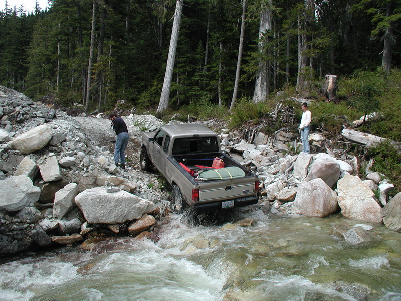 Tan Ford pickup truck driving through a rocky stream in a forest.
