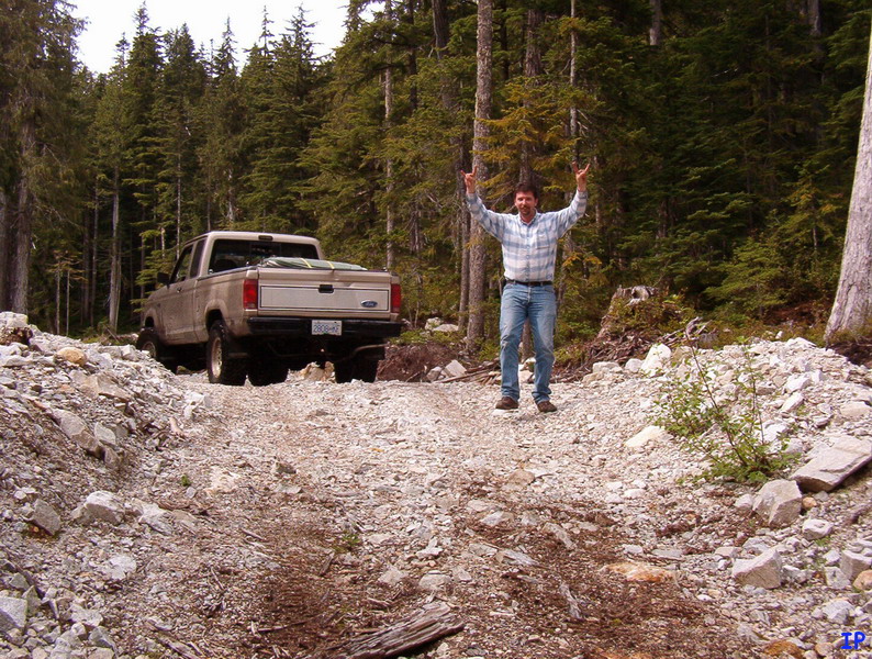 Man on a rocky trail giving rock-on hand gesture next to a tan Ford pickup truck.