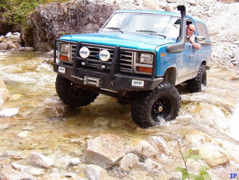 Blue 1980s Ford Bronco driving through a rocky river with off-road tires, snorkel, winch, and IPF lights.
