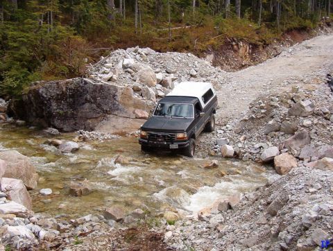 Dark pickup truck with white camper shell driving through a rocky stream on a dirt road.