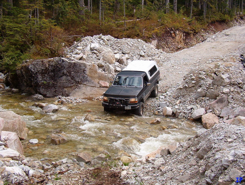 Dark pickup truck with white camper shell driving through a rocky stream on a dirt road.