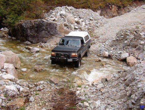 Dark pickup truck with white camper shell driving through a rocky stream.