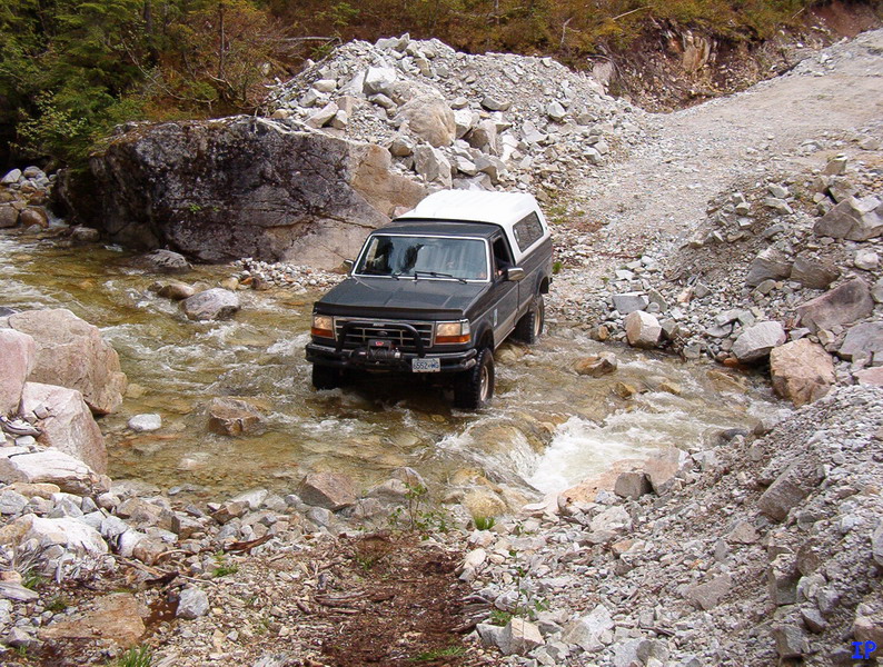 Dark pickup truck with white camper shell driving through a rocky stream.