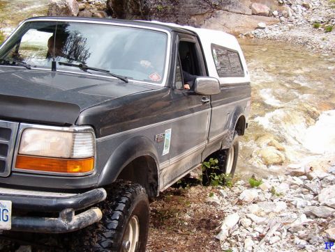 Dark gray Ford F-150 pickup truck with white camper shell driving through a rocky stream.