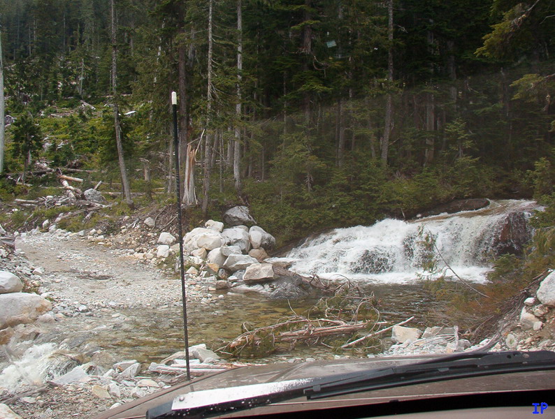 Front view of a vehicle on a gravel road near a waterfall in a forest.