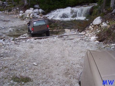 Brown SUV crossing a stream with a waterfall behind it. Another vehicle in foreground.