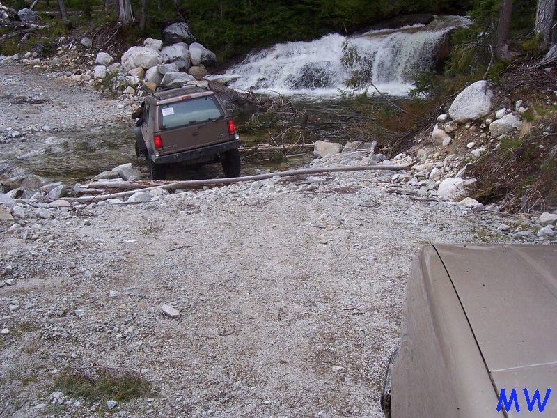 Brown SUV crossing a stream with a waterfall behind it. Another vehicle in foreground.