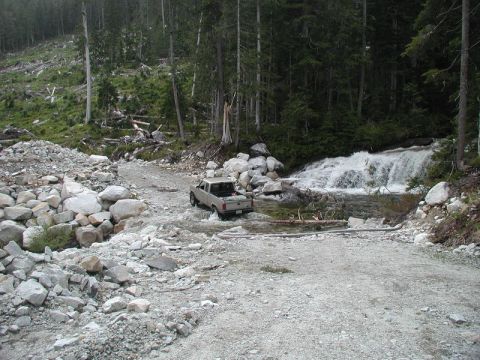 Pickup truck driving through a stream on a rocky road with a waterfall and forest.