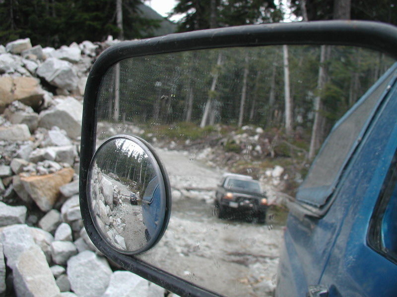 Reflection in side mirror of a black SUV on a dirt road with rocky, forested surroundings.