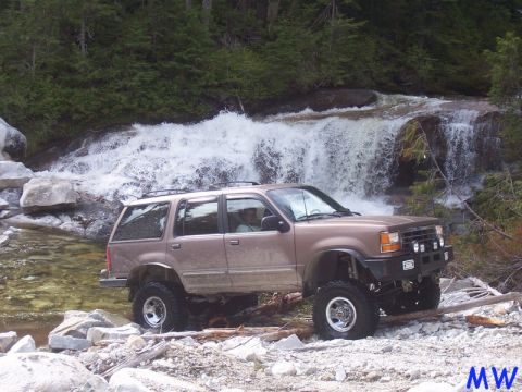 Lifted tan SUV driving through a stream near a waterfall.