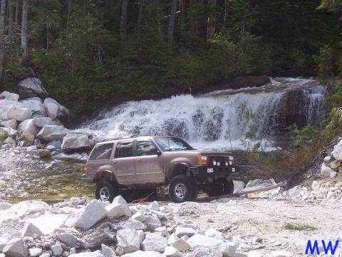 Tan Ford Explorer SUV with off-road modifications parked near a waterfall.