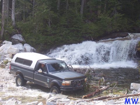 Dark gray Ford pickup truck with white camper shell driving through a river in front of a waterfall.