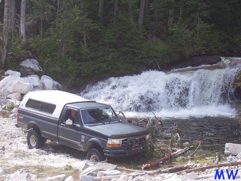 Dark gray Ford pickup truck with white camper shell driving through a river in front of a waterfall.